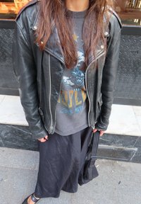 Svart skinn motorsykkeljakke med sølvfarge detaljer, grå grafisk t-skjorte, flytende svart skjørt og svarte leiligheter. Teksturerte stoffdetaljer synlige.