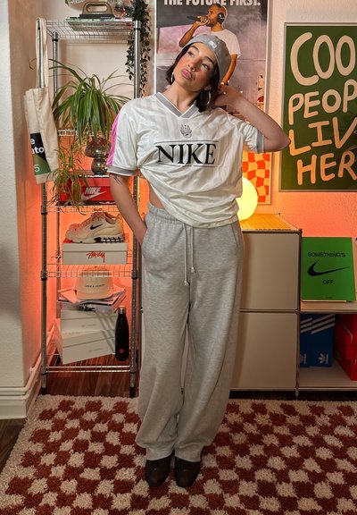 Camiseta blanca de Nike con logo negro y detalles en rosa, combinada con pantalones deportivos gris claro. Zapatos oscuros completan el look deportivo. El fondo incluye plantas y estantes.