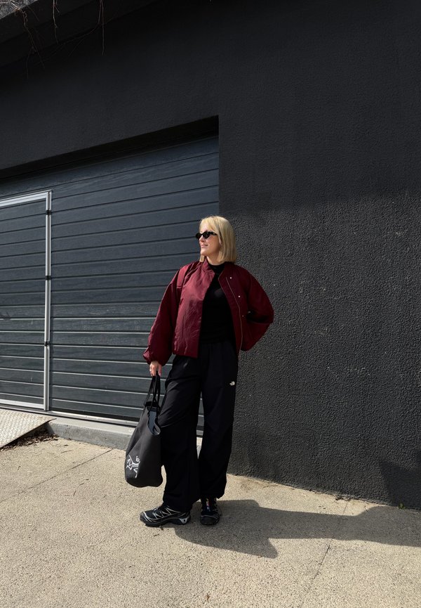 Blonde Person mit Sonnenbrille, roter Jacke, schwarzen Hosen und Schuhen steht und hält eine schwarze Tragetasche bei einer dunkelgrauen Wand und Garagentor.