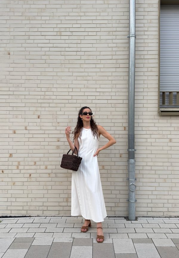 Femme en robe blanche sans manches et lunettes de soleil tenant un sac tressé marron, debout contre un mur en briques claires près d'une gouttière en métal.