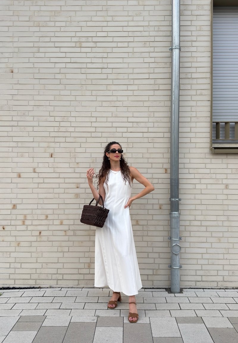 Woman in a white sleeveless dress and sunglasses holding a brown woven bag, standing against a light brick wall near a metal drainpipe.
