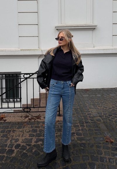 Mujer rubia con gafas de sol negras y chaqueta, jeans azules con tachuelas y botas negras, de pie en una calle empedrada con un fondo de edificio blanco.