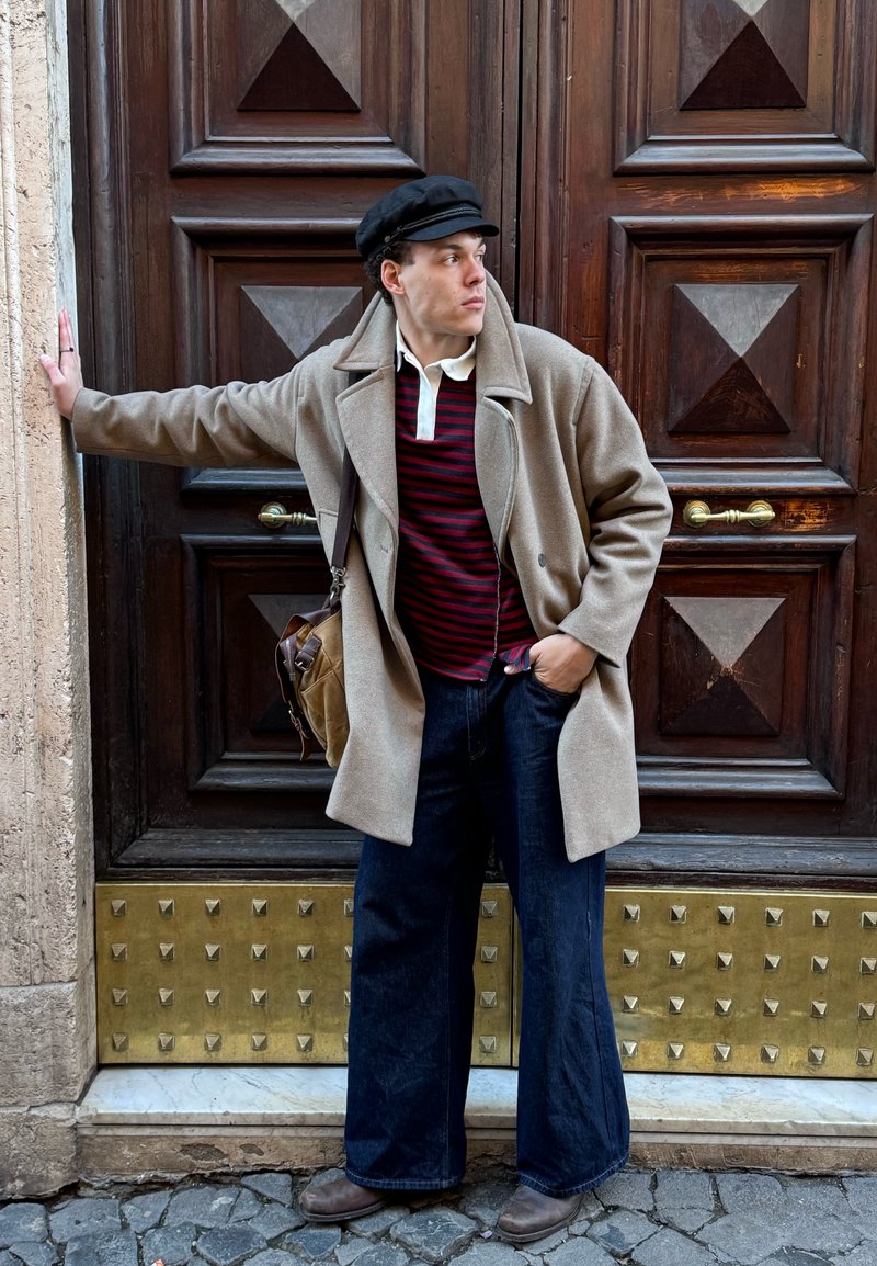 Jeune homme en manteau beige, chemise rayée, jean large et casquette noire, appuyé contre une grande porte en bois décorative avec des détails en laiton.