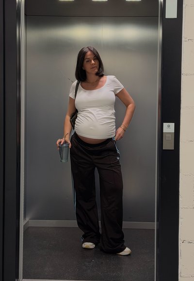 Mujer embarazada con blusa blanca y pantalones negros sosteniendo una bebida, de pie dentro de un ascensor moderno con paredes metálicas.