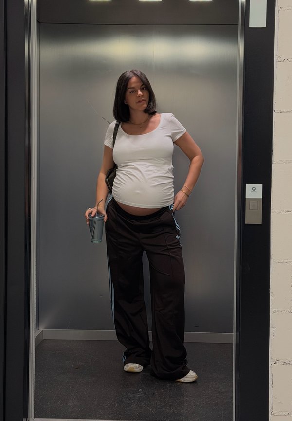 Femme enceinte en haut blanc et pantalon noir tenant une boisson, debout à l'intérieur d'un ascenseur moderne aux parois métalliques.