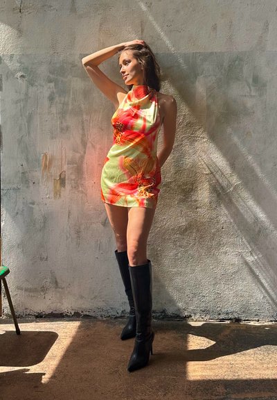 Vestido mini floral con base amarilla y patrones florales rojos y naranjas, combinado con botas de tacón negras hasta la rodilla, sobre un fondo de pared texturizada.