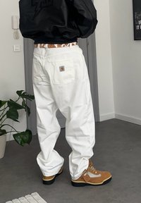 Pantalon de travail blanc ample avec une poche arrière ornée d'un logo, associé à des bottes de randonnée couleur camel avec des accents dorés et une ceinture à motifs.