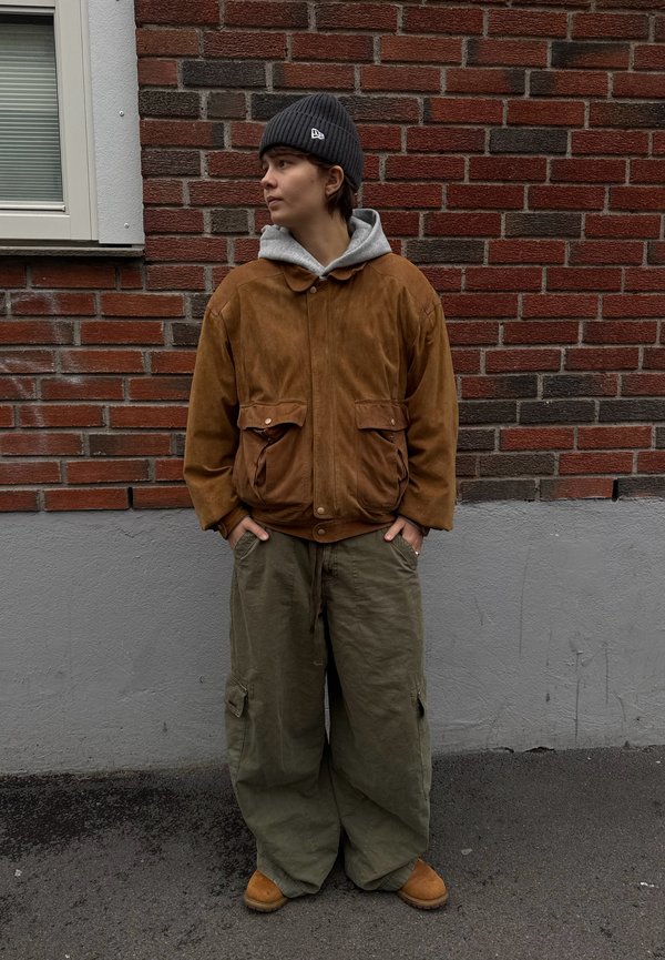 Braune Wildlederjacke über einem grauen Kapuzenpullover, kombiniert mit olivgrünen weiten Hosen und braunen Stiefeln. Im Hintergrund ist eine graue Wand mit Ziegelsteinen zu sehen.