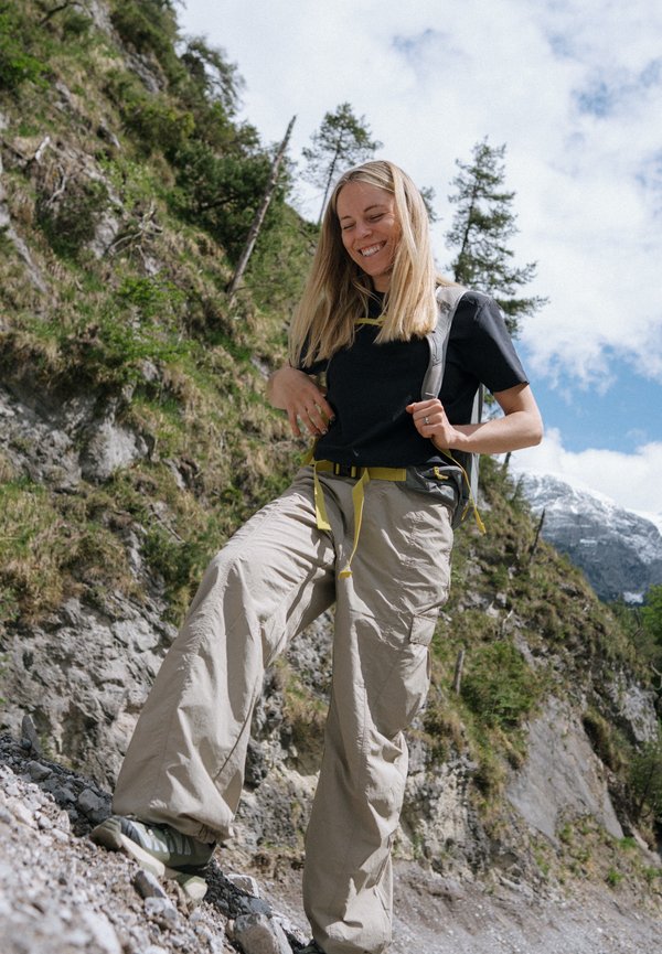 Eine Frau wandert auf einem felsigen Bergpfad, trägt beigen Hosen, ein schwarzes Shirt und einen Rucksack, lächelt mit Bäumen und Bergen im Hintergrund.