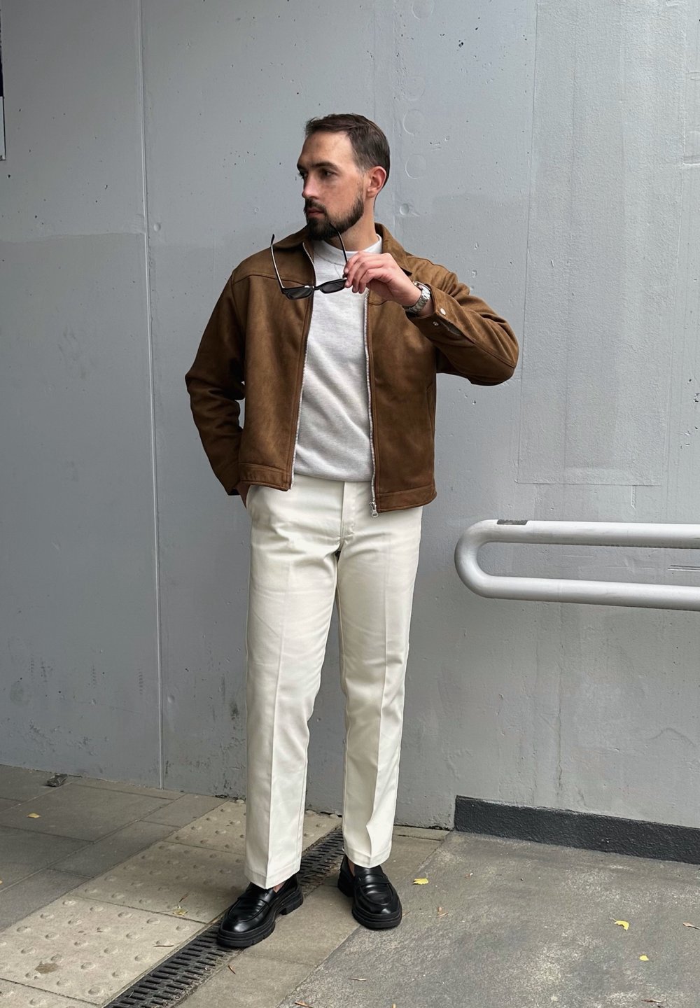 Καφέ σουέτ μπόμμπερ τζάκετ πάνω από ένα ανοιχτό γκρίζο πουλόβερ, σε συνδυασμό με λευκά παντελόνια και μαύρες λοφάδες. Το άτομο κρατά γυαλιά ηλίου. Σαφής φόντο από σκυρόδεμα.