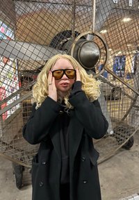 Manteau noir par-dessus un sweat à capuche foncé, cheveux blonds longs et ondulés avec des tresses, et lunettes de soleil ambrées surdimensionnées. Arrière-plan industriel avec un grand ventilateur.