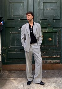 Sivý dvojradový oblek so širokými nohavicami, doplnený o čiernu polokošeľu. Lesklé slnečné okuliare; na pozadí zelených textúrovaných dverí.