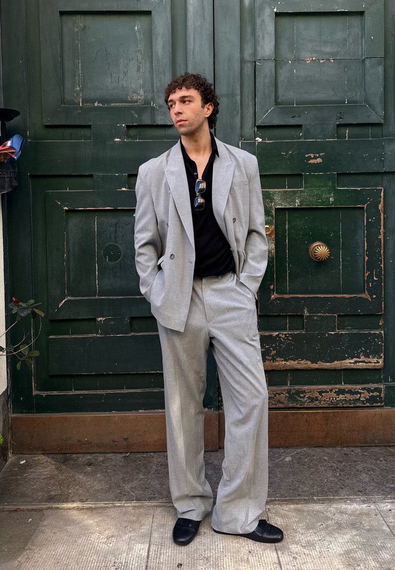 Abito grigio doppiopetto con pantaloni a gamba larga, abbinato a una polo nera. Occhiali da sole lucidi indossati; sullo sfondo, una porta verde con texture.