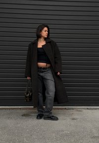 Long manteau foncé en laine, assorti à un haut noir et un jean gris. Des chaussures noires et un sac à main transparent complètent la tenue. Design simple.