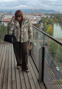Faux fur coat in cream and brown leopard print, brown pants, black handbag with decorative keychain, standing on a wooden deck. City view in background.