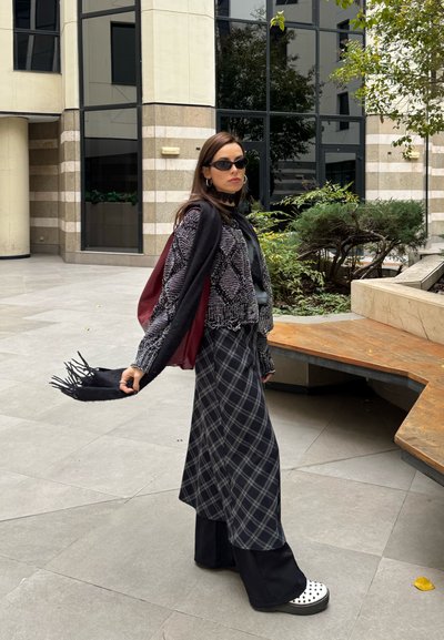 Mujer con gafas de sol, suéter con estampado, falda de cuadros, pantalones negros, zapatos blancos, sosteniendo una bufanda negra, de pie en un patio urbano con plantas y un edificio.
