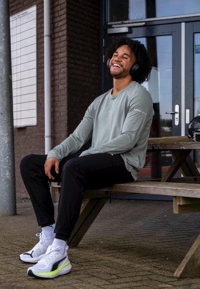 Camisa de manga larga gris claro, pantalones deportivos negros y zapatillas blancas con acentos en negro y verde neón. Persona sentada en un banco de madera.