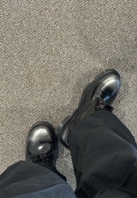 Botas de couro pretas com atacadores, usadas com calças pretas, em pé sobre um tapete cinza texturizado com um pequeno padrão xadrez.