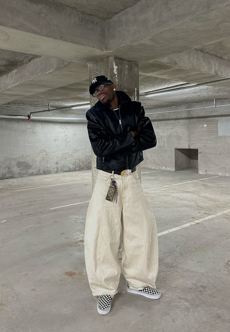 Personne portant une veste noire, un pantalon large blanc, des chaussures à enfiler à carreaux et une casquette, debout les bras croisés dans un parking en béton.