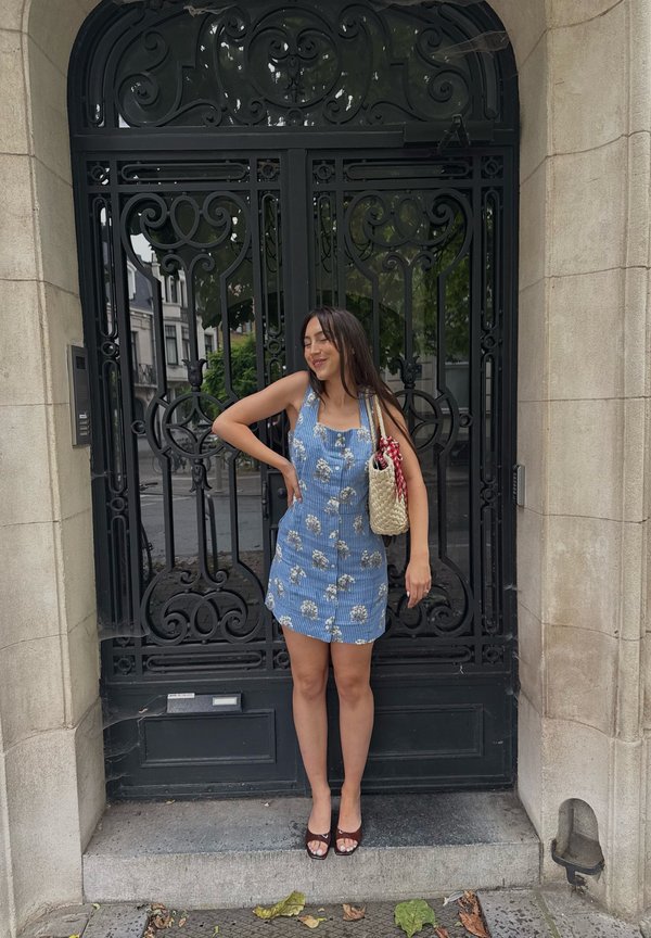 Donna in abito blu a fiori, sorridente con una mano sui fianchi davanti a un cancello nero in ferro battuto decorato.
