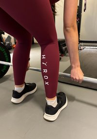 Personne en leggings bordeaux et baskets noires tenant une barre au sol de la salle de sport, se préparant pour un soulevé de terre ou un exercice de musculation.