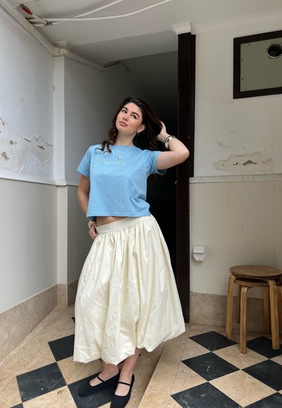 Mujer con cabello largo castaño, vistiendo una camiseta corta azul, una falda midi color crema y zapatos planos negros, de pie sobre un suelo de cuadros en interiores.