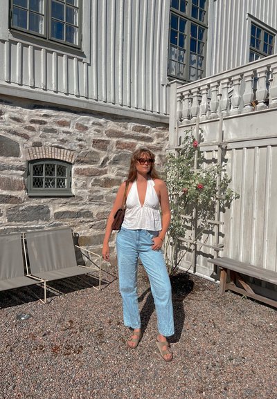 Top halter blanco, jeans rectos azul claro, sandalias beige; de pie sobre grava con un muro de piedra y estructuras de madera de fondo.
