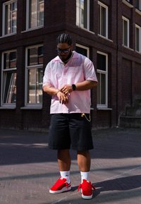 Homem com cabelo trançado a usar óculos de sol, uma camisa rosa, calções pretos, meias brancas e ténis vermelhos ajusta o seu bracelete numa esquina de rua da cidade perto de edifícios de tijolo.