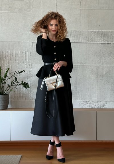 Cárdigan negro texturizado con botones, cintura de peplum, combinado con una falda larga negra. Bolso metálico dorado con correa negra y tacones negros.