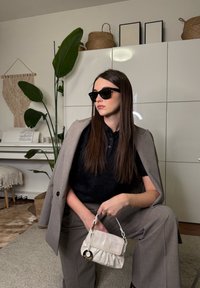 Γυναίκα με γκρι κοστούμι και μαύρο πουκάμισο που φοράει γυαλιά ηλίου μέσα σε κλειστό χώρο, κρατώντας μια μικρή λευκή τσάντα, καθισμένη σε έναν μοντέρνο σαλόνι με φυτά.