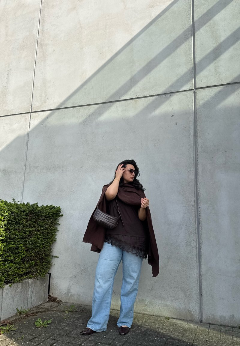 Žena s naočalama za sunce, smeđom majicom i šalom, svijetloplavim trapericama, stoji uz sivu betonsku zid s sjenom i zelenim grmom sa strane.