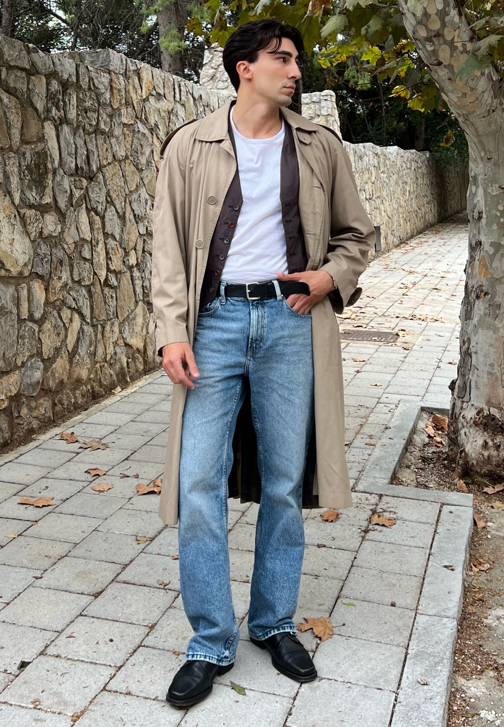 Hellbeige Trenchcoat über einem dunklen Hemd, weißem T-Shirt und hellblauer Jeans; schwarze Lederschuhe; auf einem steinernen Weg stehend.