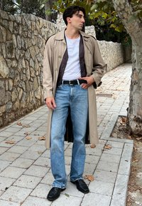 Hellbeige Trenchcoat über einem dunklen Hemd, weißem T-Shirt und hellblauer Jeans; schwarze Lederschuhe; auf einem steinernen Weg stehend.