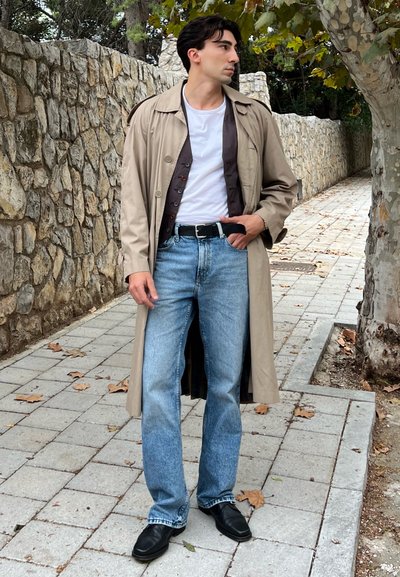 Trench coat beige claro sobre una camisa de botones oscura, camiseta blanca y jeans azules claros; zapatos de cuero negros; de pie sobre un camino de piedra.