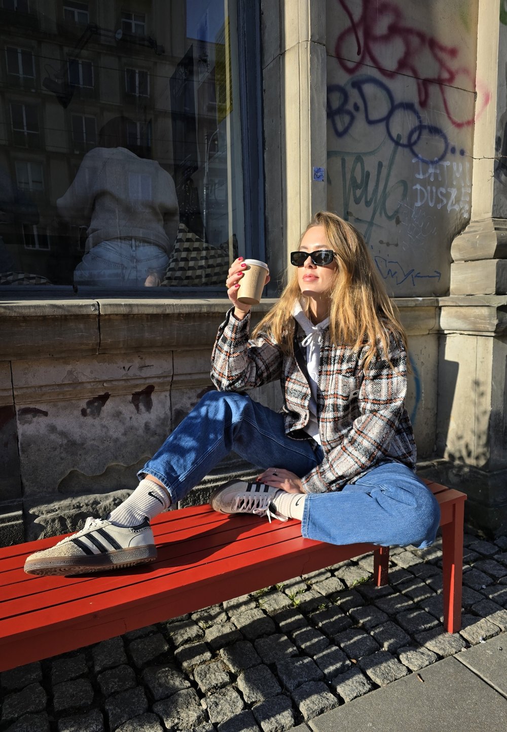 Utendørsscene med en kvinne i en rutete jakke, blå jeans og grå sneakers, sittende på en rød benk og holder en kaffekopp.