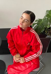Rdeč adidas trenirni komplet z belimi črtami, ki vključuje žepno jakno in usklajene hlače. Jasna steklenica z napitkom, ki jo držimo v rokah, sedi na stolu.