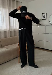 Pull en maille noire et pantalon large noir ajusté, complétés par une casquette noire avec broderie blanche et des chaussures noires. Intérieur.