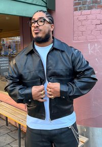 Blouson aviateur en cuir noir avec une fermeture éclair à l'avant, deux poches devant, et un col pointu, porté sur un t-shirt blanc et un pantalon noir.