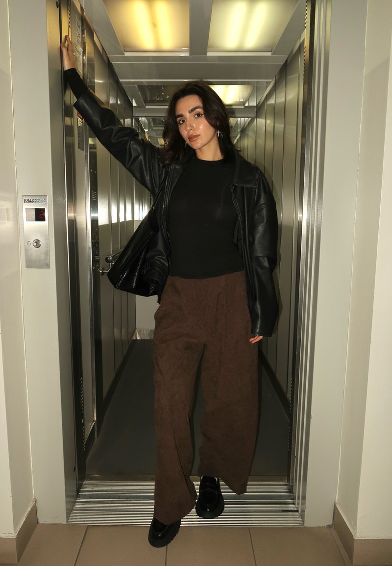 Mulher com uma jaqueta de couro preta, blusa preta e calças largas castanhas, de pé em um elevador aberto com um braço apoiado no batente da porta.