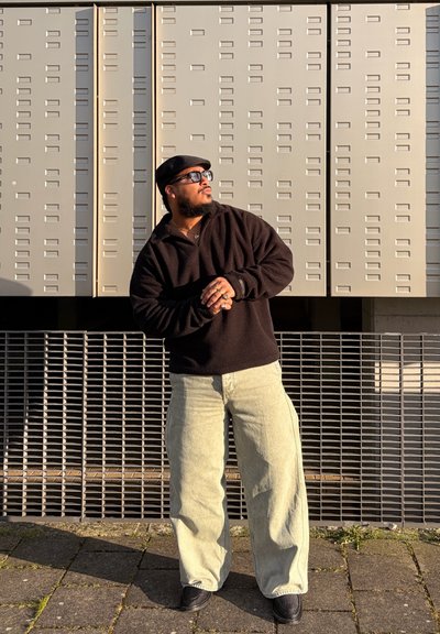 Hombre con gorra negra, gafas de sol y suéter oscuro con pantalones anchos claros, está apoyado contra una pared texturizada a la luz del sol, mirando hacia un lado.