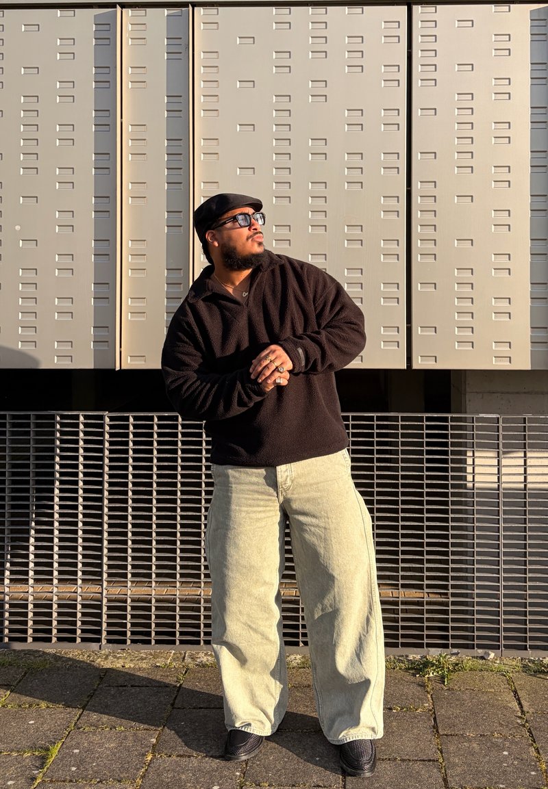 Homme portant une casquette noire, des lunettes de soleil et un pull sombre avec un pantalon large clair, debout contre un mur texturé au soleil, regardant sur le côté.
