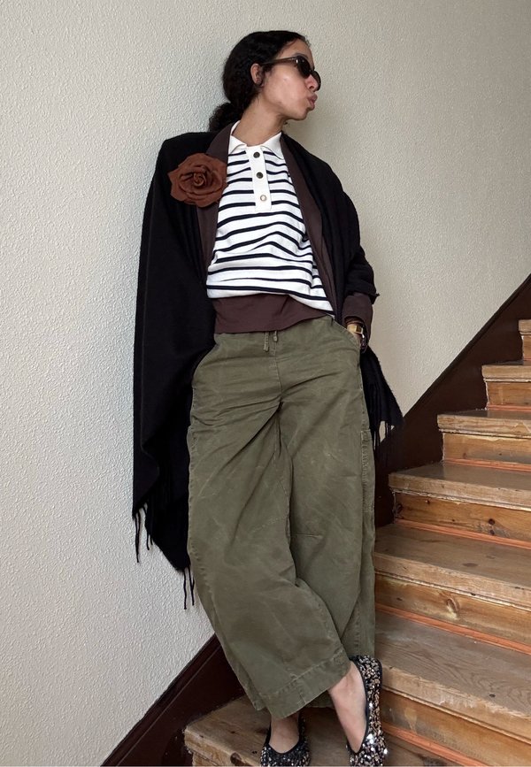 Personne portant un châle noir, une chemise rayée avec une broche fleur marron, un pantalon large olive et des ballerines à sequins, appuyée contre un mur près d'un escalier en bois.