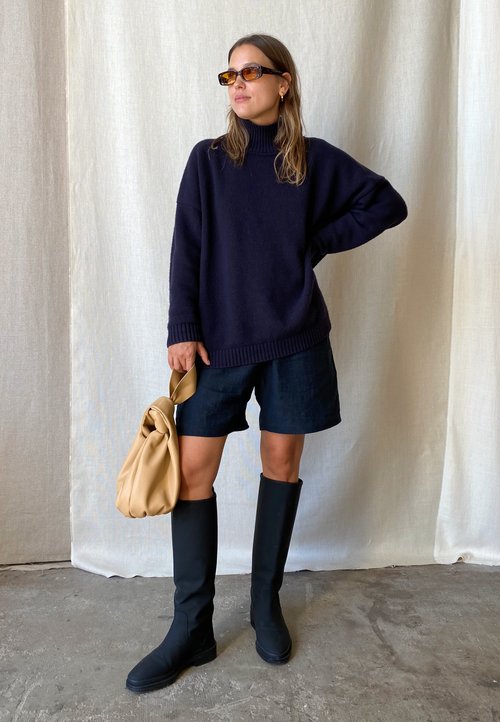 Ylipitkä laivaston sininen neule, tummat shortsit, mustat polvipituiset saappaat ja ruskea laukku, jossa on solmittu kahva. Sujuvat tekstuurit ja moderni siluetti.