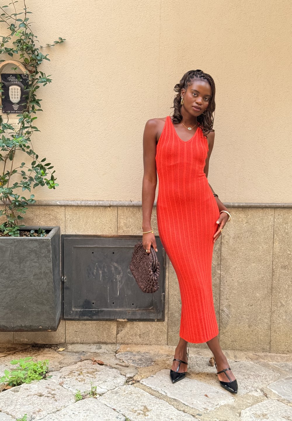 Red, knitted midi dress with a V-neck and ribbed texture. Paired with black pointed-toe heels and a woven clutch, set against a pale wall.