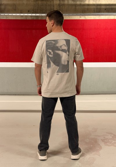 Camiseta de algodón gris claro con un gran gráfico en blanco y negro en la espalda. Combinada con pantalones gris oscuro y calzado blanco.