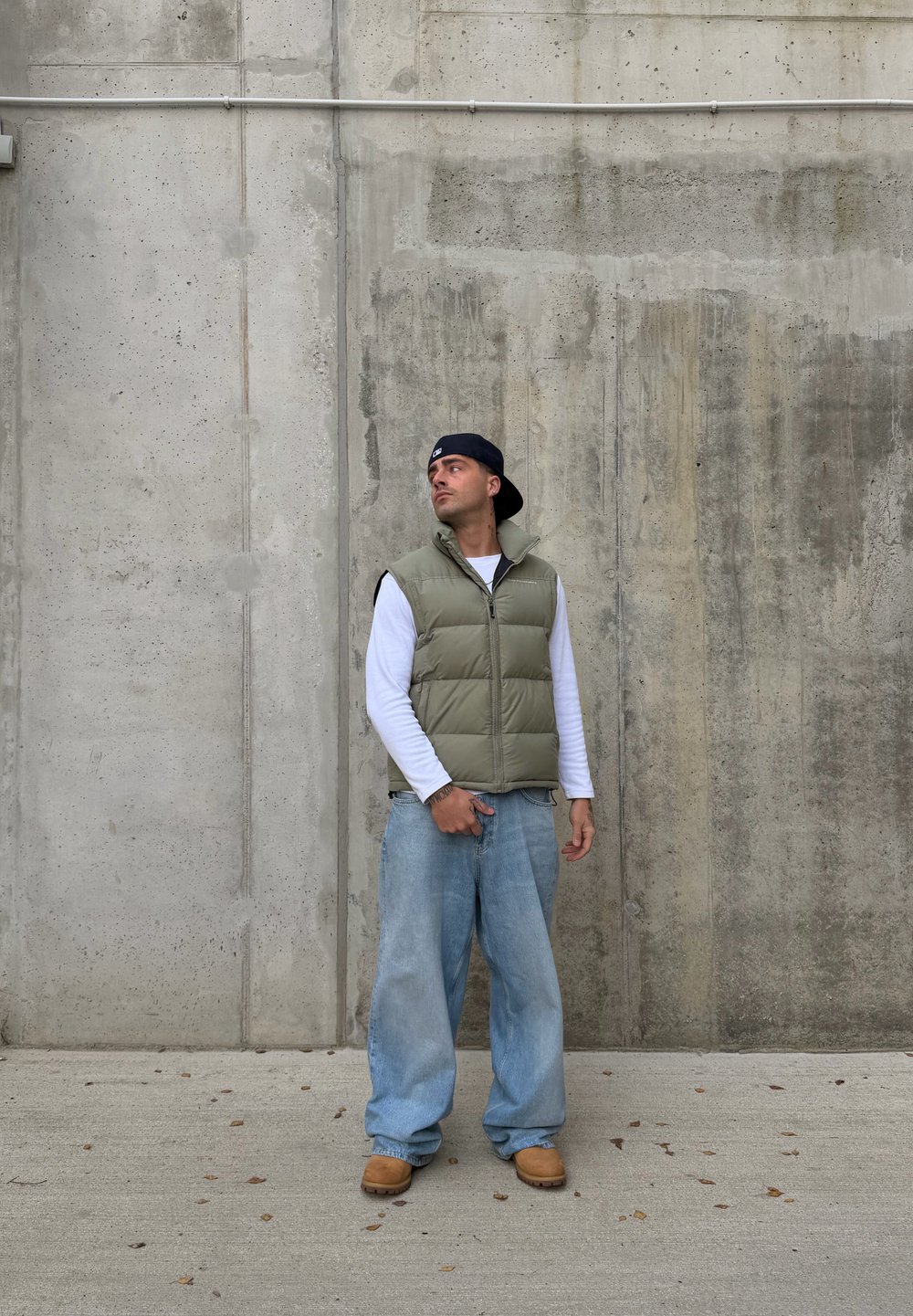 Uomo con un cappellino nero indossato al contrario, giubbotto imbottito verde, camicia bianca a maniche lunghe, jeans larghi e stivali marroni, in piedi contro un muro di cemento.