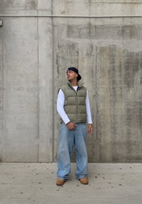 Uomo con un cappellino nero indossato al contrario, giubbotto imbottito verde, camicia bianca a maniche lunghe, jeans larghi e stivali marroni, in piedi contro un muro di cemento.