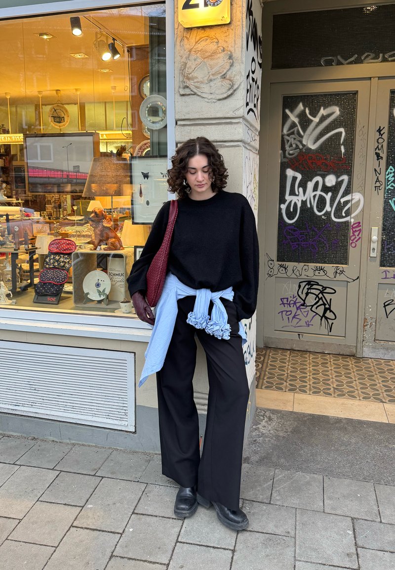 Giovane con capelli ricci che indossa un maglione nero e pantaloni, camicia blu annodata in vita, in piedi davanti a un negozio con porta coperta di graffiti.