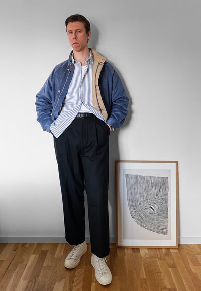 Chaqueta azul y beige sobre una camiseta de rayas azul y blanca, pantalones negros y zapatillas blancas. Obra de arte abstracto enmarcada al fondo.