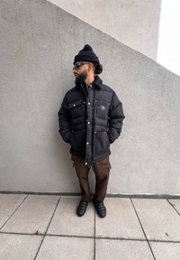 Μαύρο φουσκωτό μπουφάν με γιακά, τέσσερις μπροστινές τσέπες και ασημένιες λεπτομέρειες. Καφέ cargo παντελόνι και μαύρες παντόφλες. Γκρι φόντο.